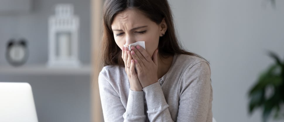 A young brunette woman sits at home and blows her nose, looking as if she is in pain.