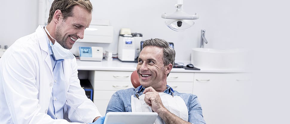 Patient in a dental chair smiles at a dentist as he is shown something on the dentist’s tablet