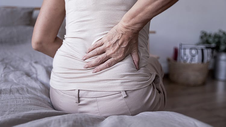 A woman sits on her bed holding her back in pain