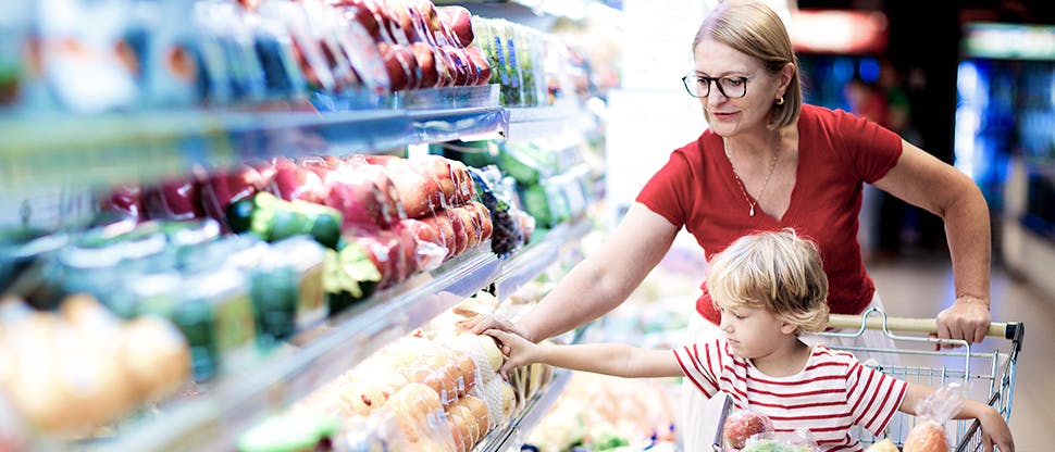 A woman pushes a shopping trolley with a child in and they both reach out to select items from a shelf.