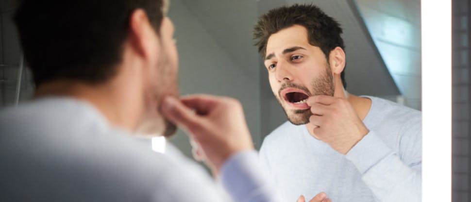 A man examines his inner mouth in the bathroom mirror searching for signs of gingivitis or gum disease.