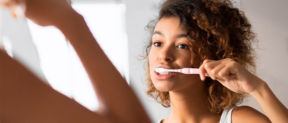 A young woman brushes her teeth in the bathroom mirror with a toothbrush.