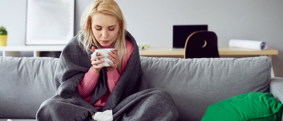 Woman sitting on a sofa wrapped in a blanket blows on her coffee