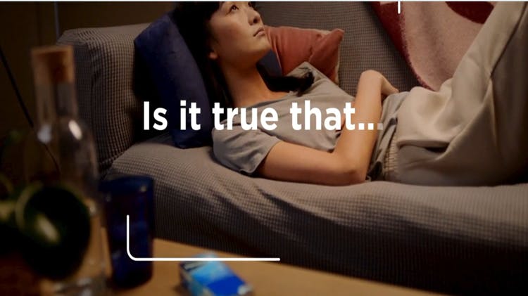 A brunette woman lies on the couch, possibly experiencing headache or body pain, while a Panadol product sits on the coffee table in the foreground.