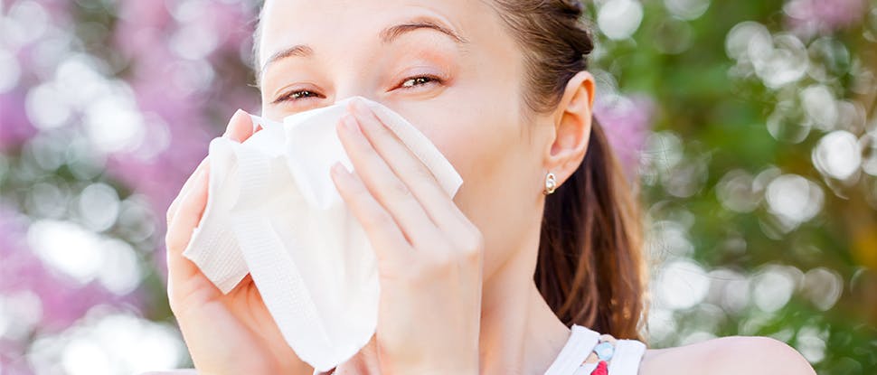 A young brunette woman blows her nose as she stands outside. The background is blurred but looks like blossom trees.