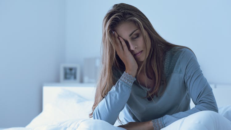 Young brunette woman sits up in bed and looks tired while holding her head in one hand