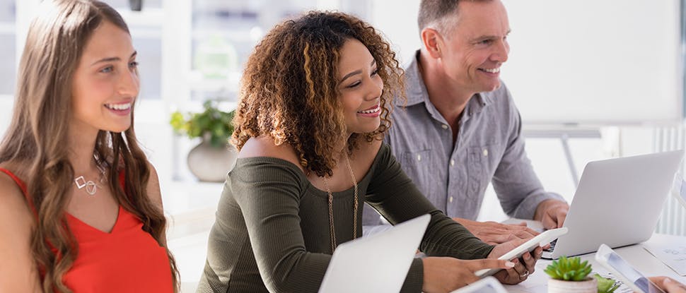 Three smiling colleagues – two younger women and one older man – sit in front of laptops while collaborating at work 