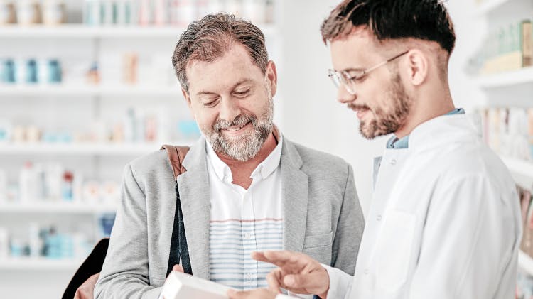 Younger male healthcare professional wearing glasses speaks to an older male patient about medication in a pharmacy