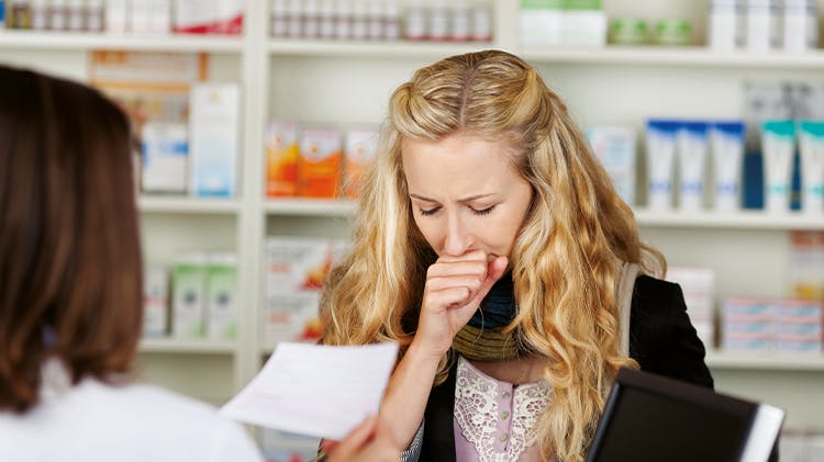 Young woman with long wavy blonde hair stands in front of a female brunette pharmacist in a pharmacy coughing