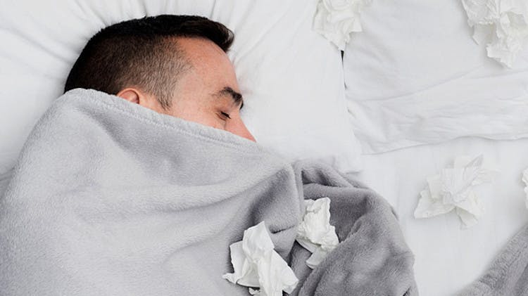 Man covered in a blanket on the bed with some tissues on his blanket