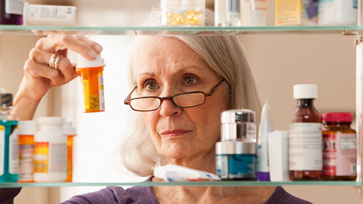 An older blonde woman wearing glasses inspects a pill receptacle in a medicine cabinet.