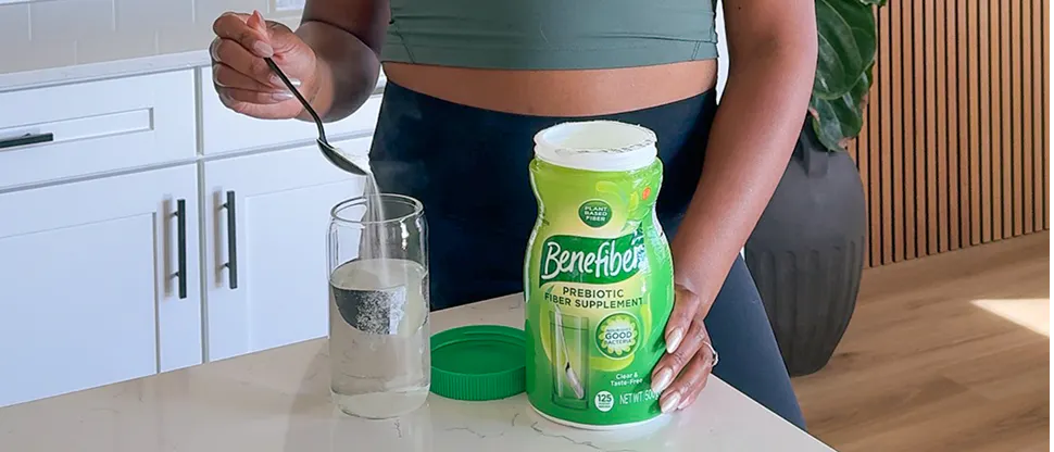 Person standing at a kitchen counter preparing a drink with a spoon and a glass of water next to an open container of Benefiber.