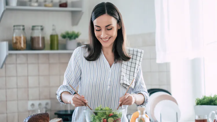 A person standing in a kitchen preparing a bowl of salad at a counter with various fruits, vegetables, and kitchen items around them.