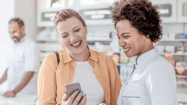 Two people standing together in a pharmacy looking at a smartphone, with shelves of products in the background and another person standing behind the counter.
