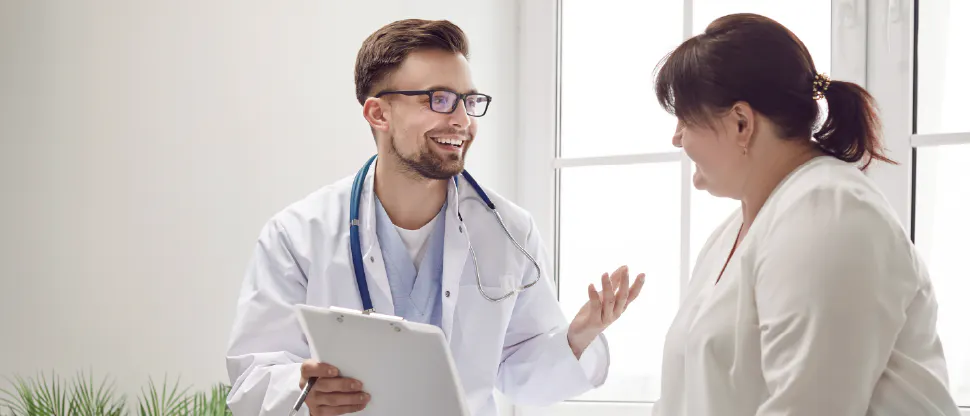 A doctor holding a clipboard sits across from another person in a bright room with large windows.