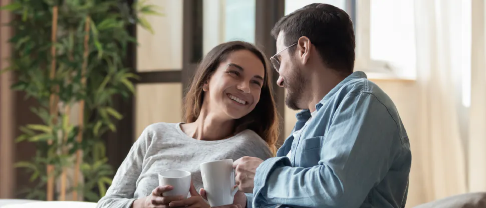 Two people sitting on a couch holding mugs and facing each other in a living room with large windows and a tall plant in the background
