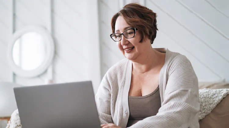 A woman sitting indoors on a sofa using a laptop, with light-colored walls in the background.