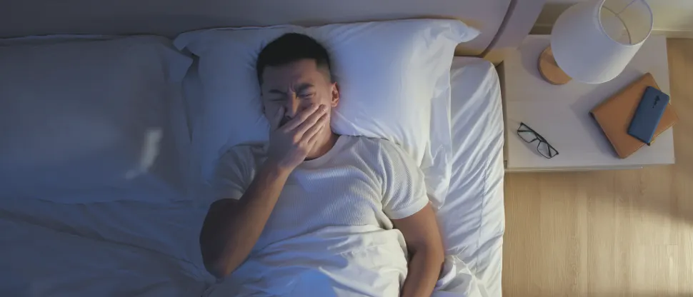 A person lies on a bed under white bedding, wearing a white T‑shirt, with one hand covering the mouth