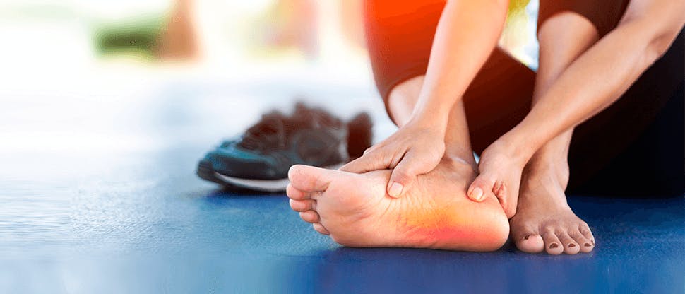 A woman with bare feet wearing exercise clothes who is clutching her foot with a highlight to show pain in the sole from plantar faciitis