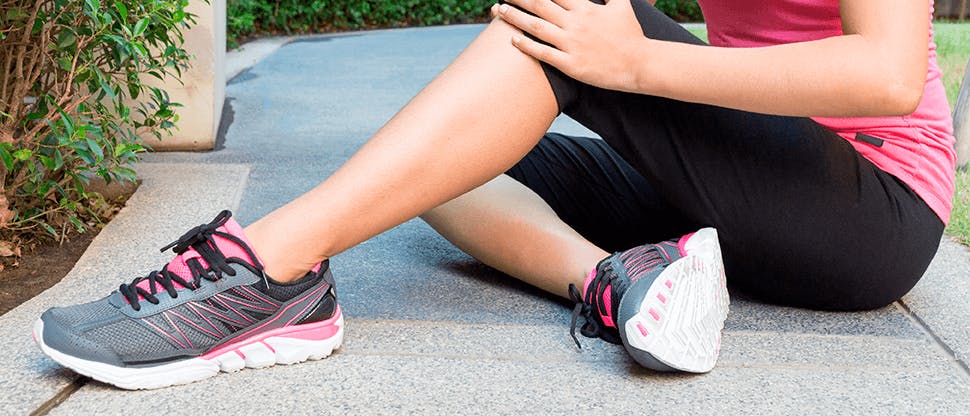 A woman in exercise leggings and sneakers clutches her knee while seated in the ground outside as a result of pain from arthritis.