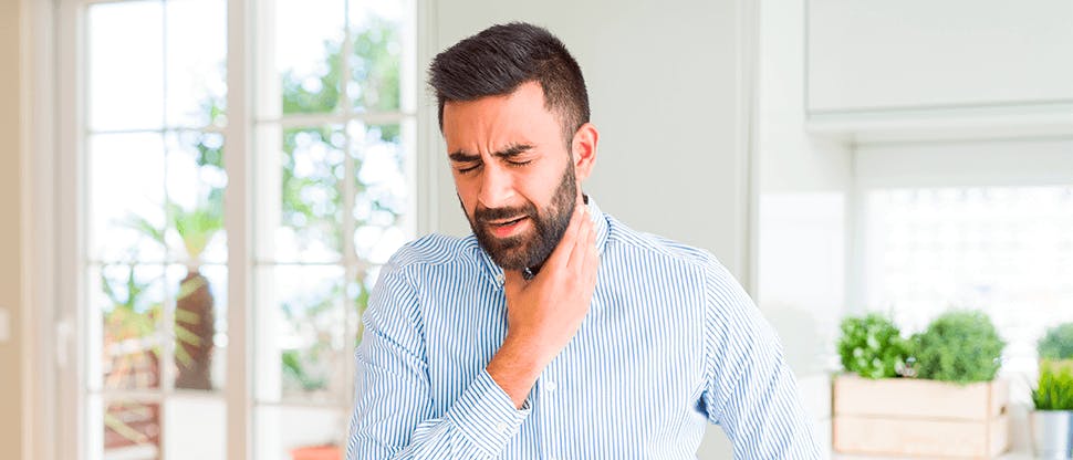 A man wearing a striped shirt stood in his home clutches the sides of his neck as an indication of throat pain from pharyngitis.