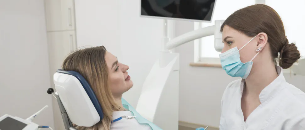 A woman in a white shirt sits in a dentist chair while a healthcare professional in scrubs speaks to them