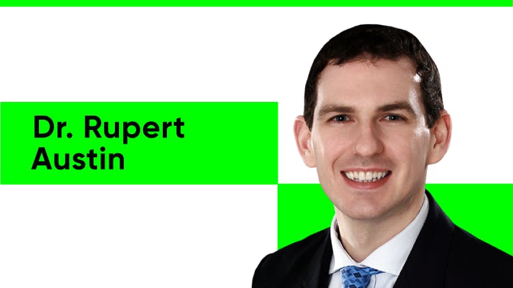 Portrait du Dr Rupert Austin, un homme blanc aux cheveux noirs courts. Il sourit tout en portant une chemise blanche, un blazer noir et une cravate bleue à motifs