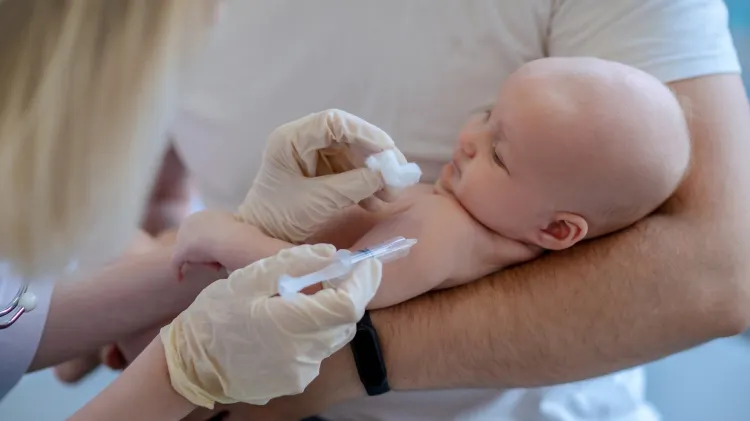 Egy apa tartja csecsemőjét, miközben egy egészségügyi szakember vakcinát ad be a felkarjába