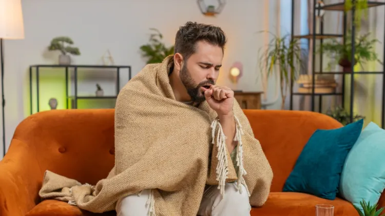 Fiatal férfi ül a nappalijában megfázással küzdve, takaróba burkolózva köhög, miközben a kezét a szájához emeli