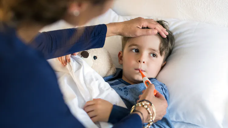 Ágyban fekvő kisgyermeknek szájában lázmérő. Egy fiatal nő fölé hajol, egyik keze a gyermek homlokán, másik kezével a hőmérőt fogja