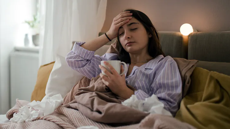 Egy influenzás tüneteket mutató fiatal nő fekszik az ágyban, kezében egy bögre forró itallal, körülötte használt zsebkendők