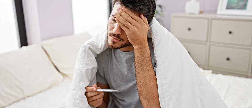 Egy paplanba burkolózott férfi ül az ágyban, a fejét fogva, miközben a hőmérőt nézi
