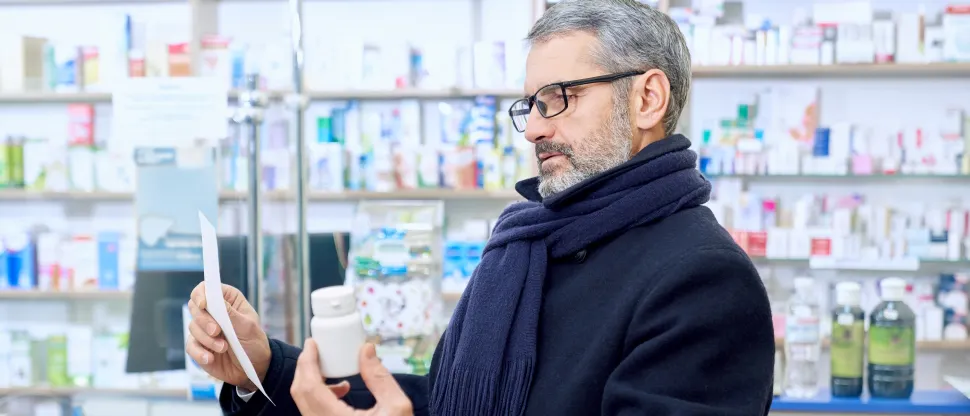 Középkorú férfi a gyógyszertárban kezében receptet és gyógyszeres dobozt tart, és a gyógyszerrel kapcsolatos információkat olvassa