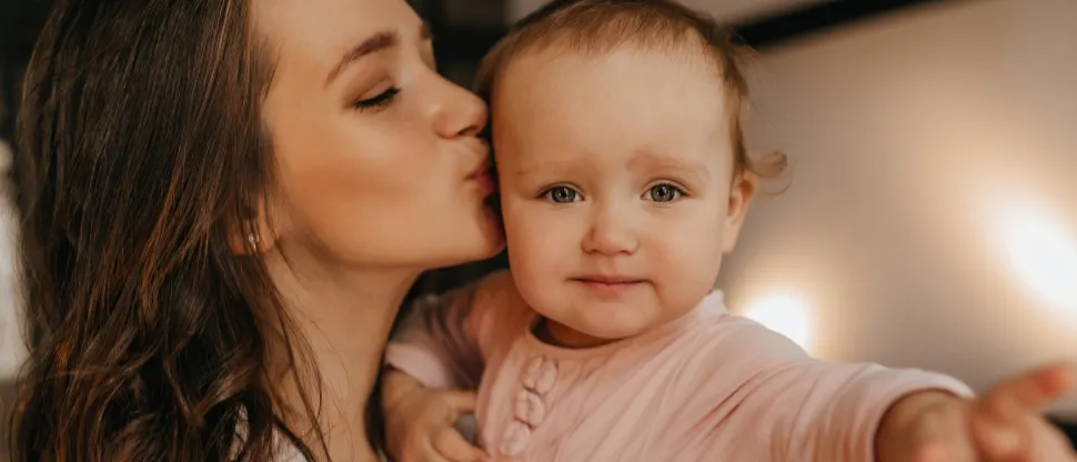 Fiatal édesanya karjában tartja és megcsókolja mosolygó kisbabáját. Mindkettőjüknek barna haja van