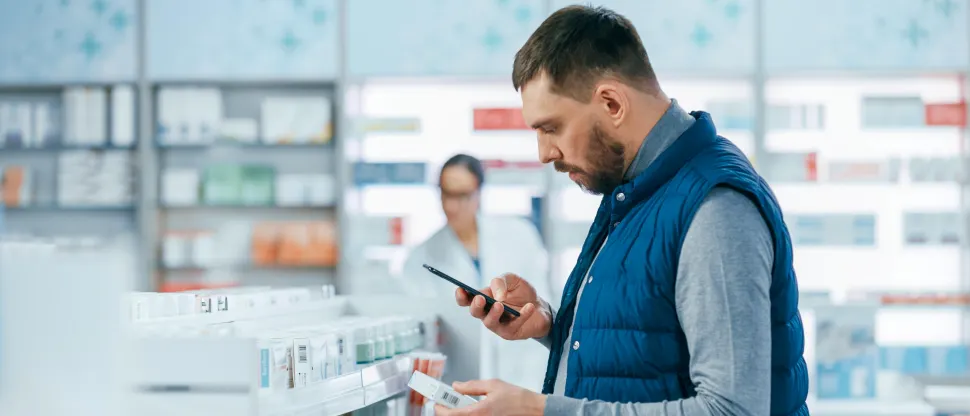 Fiatal férfi a gyógyszertárban gyógyszeres dobozt tart a kezében, miközben telefonja kijelzőjét nézi