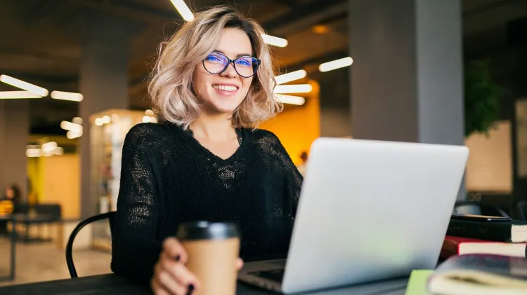 Mosolygó, szőke göndör hajú fiatal nő sötét színű felsőben dolgozik laptopján az íróasztalánál. Mellette az asztalon egy műanyag kávéspohár, egy toll és könyvek láthatók