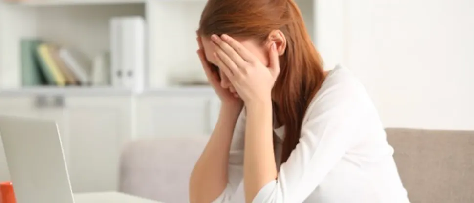 Stresszes beteg – vörösesbarna hajú fiatal nő – ül az íróasztalnál laptopja előtt, miközben ujjait a halántékához szorítja