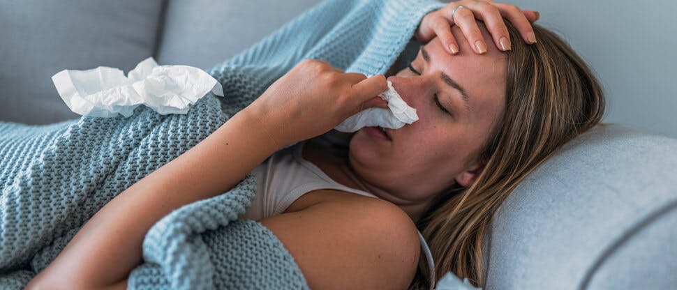 Femeie care suferă de febră, culcată pe pat, acoperită cu o pătură și care își șterge nasul cu o batistă