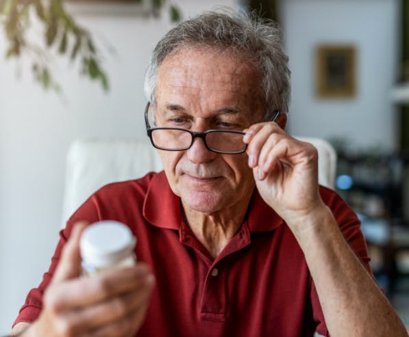 Een oudere man met bril leest de verpakking van medicijnen terwijl hij zijn bril vast houdt met zijn vrije hand
