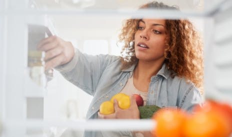 Een vrouw staat voor een open koelkast met fruit in haar handen