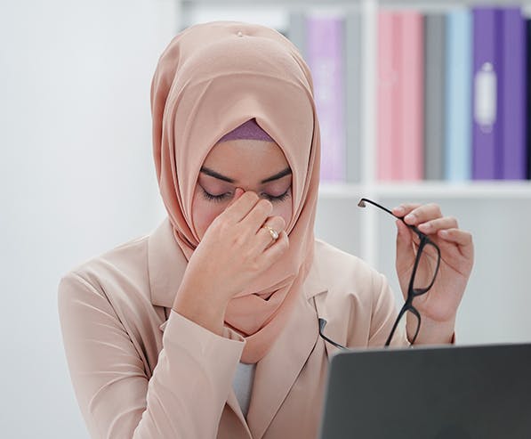 Een vrouw met een hijab zit achter een laptop en houdt haar bril vast met een hand terwijl ze met haar andere hand over haar neus wrijft