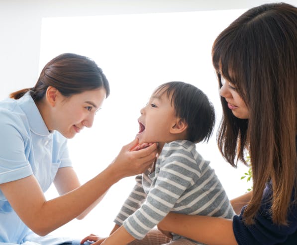 Een vrouw houdt een kind vast terwijl een arts zijn mond open houdt en in zijn keel kijkt