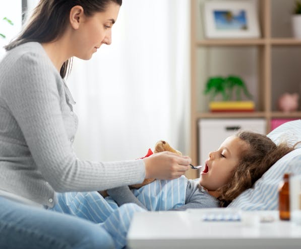 Een vrouw zit op de rand van een bed waarin een kind ligt en geeft het kind medicijnen met een lepel