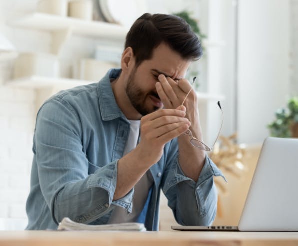 Een man zit aan een bureau voor een laptop, houdt zijn bril vast met één hand en drukt met de andere op zijn ogen