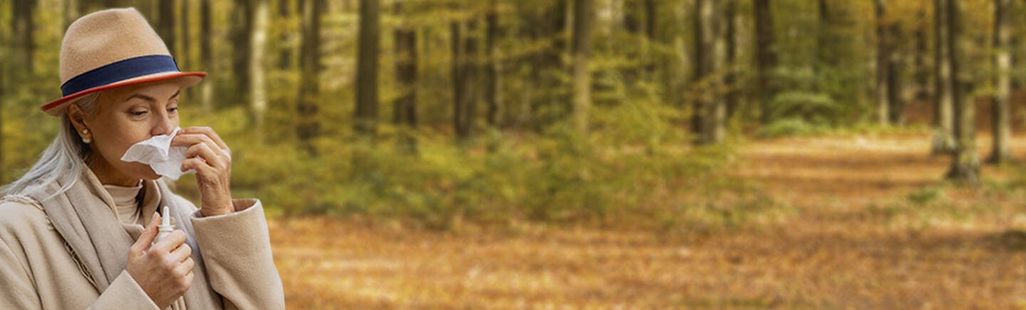 Older woman clearing her nose of air pollution as she avoids cars and traffic on the other side of a small wooded area.