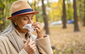 Older woman clearing her nose of air pollution as she avoids cars and traffic on the other side of a small wooded area.