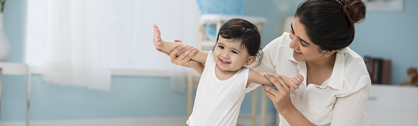 A toddler appears to smile as he stands with the support of her smiling mother