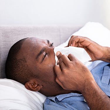 Man, with a dog next to him, is suffering from cold symptons as blocked or runny nose and he's using a tissue.