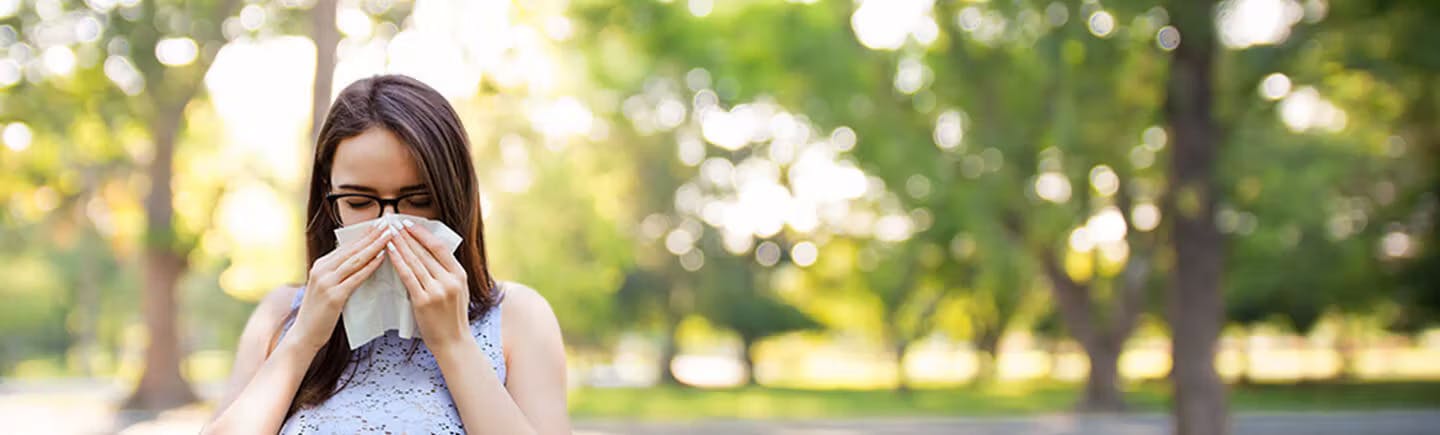 Young woman points to her nose, an organ of the respiratory system that helps you breathe by filtering impurities from the air. Kvinne med mørkt hår står ute og snyter seg i et papirlommetørkle grunnet allergi