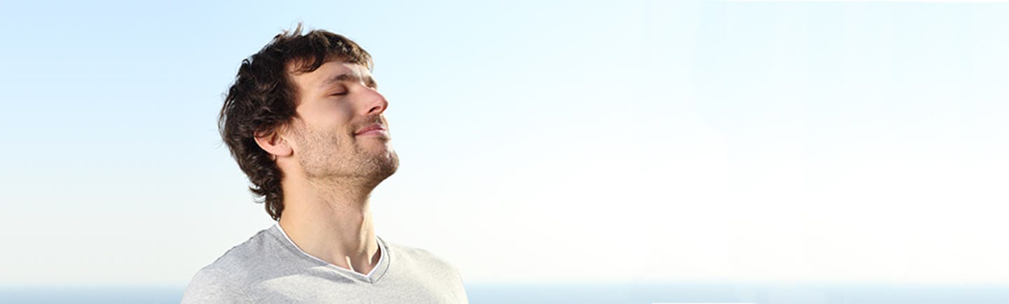 Mann med brunt hår står ute i blå himmel og puster i frisk luft med lukkede øyne